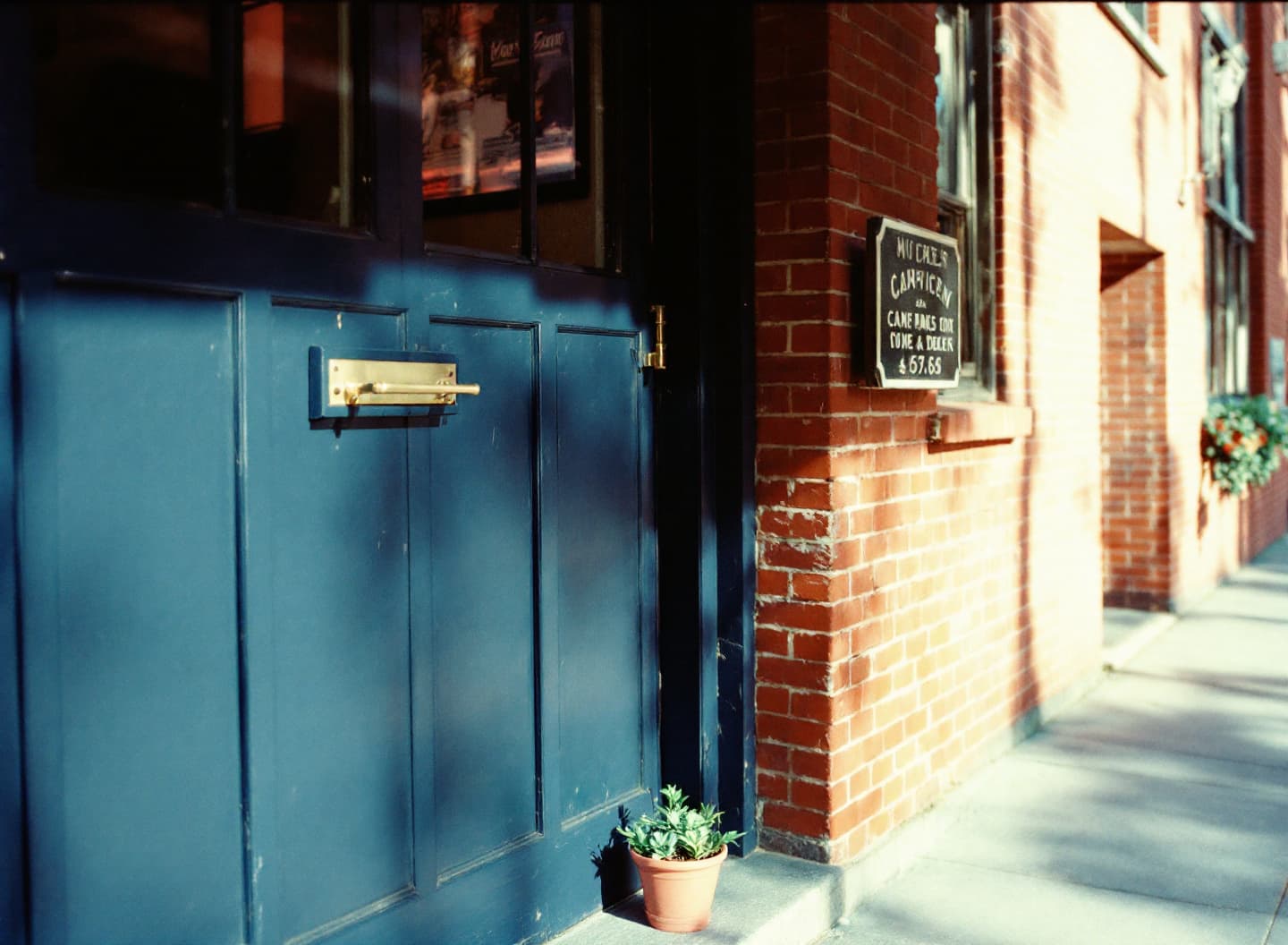 The open wooden front door of a small shop in a red brick building, with potted plants and a bench beside it